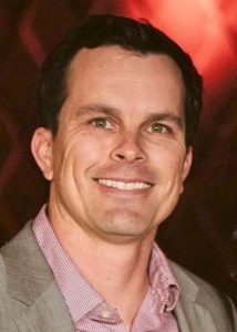 A man with short dark hair, wearing a light grey blazer over a red and white checkered shirt, smiles at the camera. The background is dimly lit with warm, reddish tones, capturing the inviting ambiance often seen in Berkeley Journalism settings.