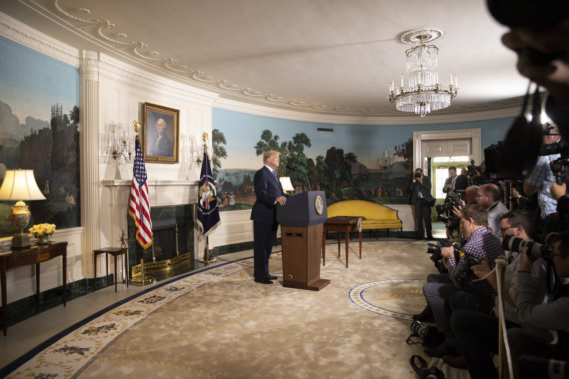 A prominent figure is giving a speech at a podium in an elegantly decorated room with chandeliers, a fireplace, and wall paintings. Flags stand behind the speaker as an audience of photographers and reporters, many of whom are from Berkeley Journalism, capture the event.