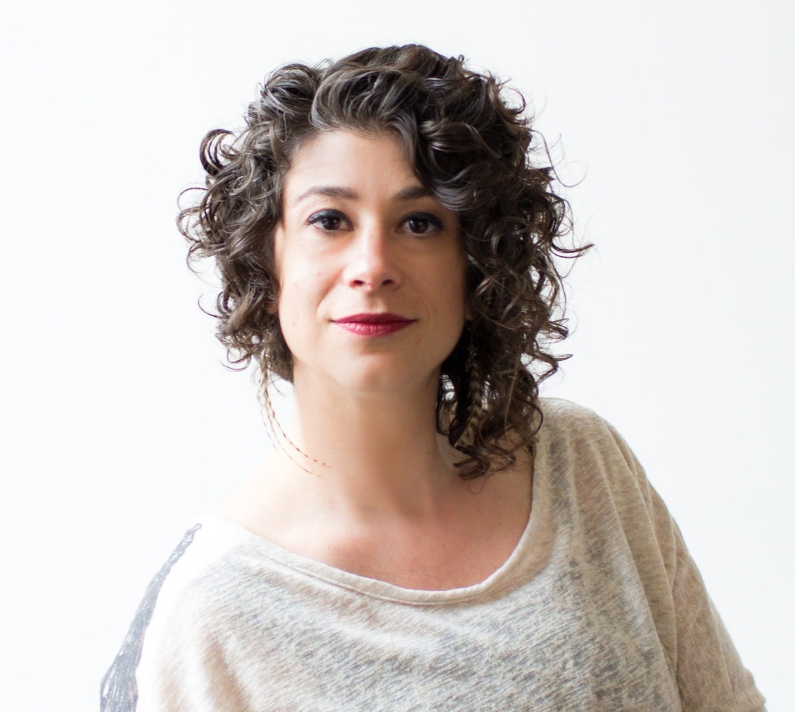 A person with short, curly dark hair is smiling slightly at the camera. They are wearing a light-colored, loose-fitting top. The background is plain and white, creating a clean and simple look, reminiscent of the candid yet polished portraits often seen in Berkeley Journalism profiles.