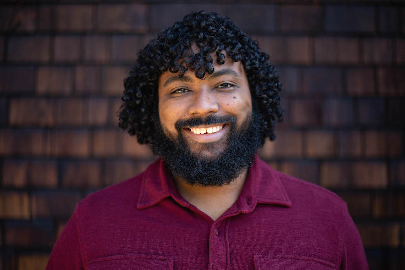 A person with curly hair and a beard smiles warmly at the camera. Wearing a red shirt and standing in front of a dark brown, shingled wall, they exude the thoughtful charm often associated with Berkeley Journalism graduates.