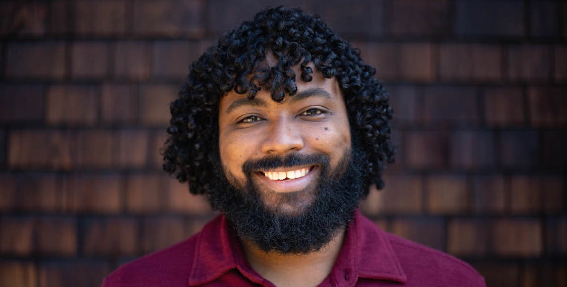 A person with curly hair and a beard, likely a Berkeley Journalism student, is smiling in front of a dark brown shingle wall. They are wearing a maroon shirt. The background and lighting are natural and warm.