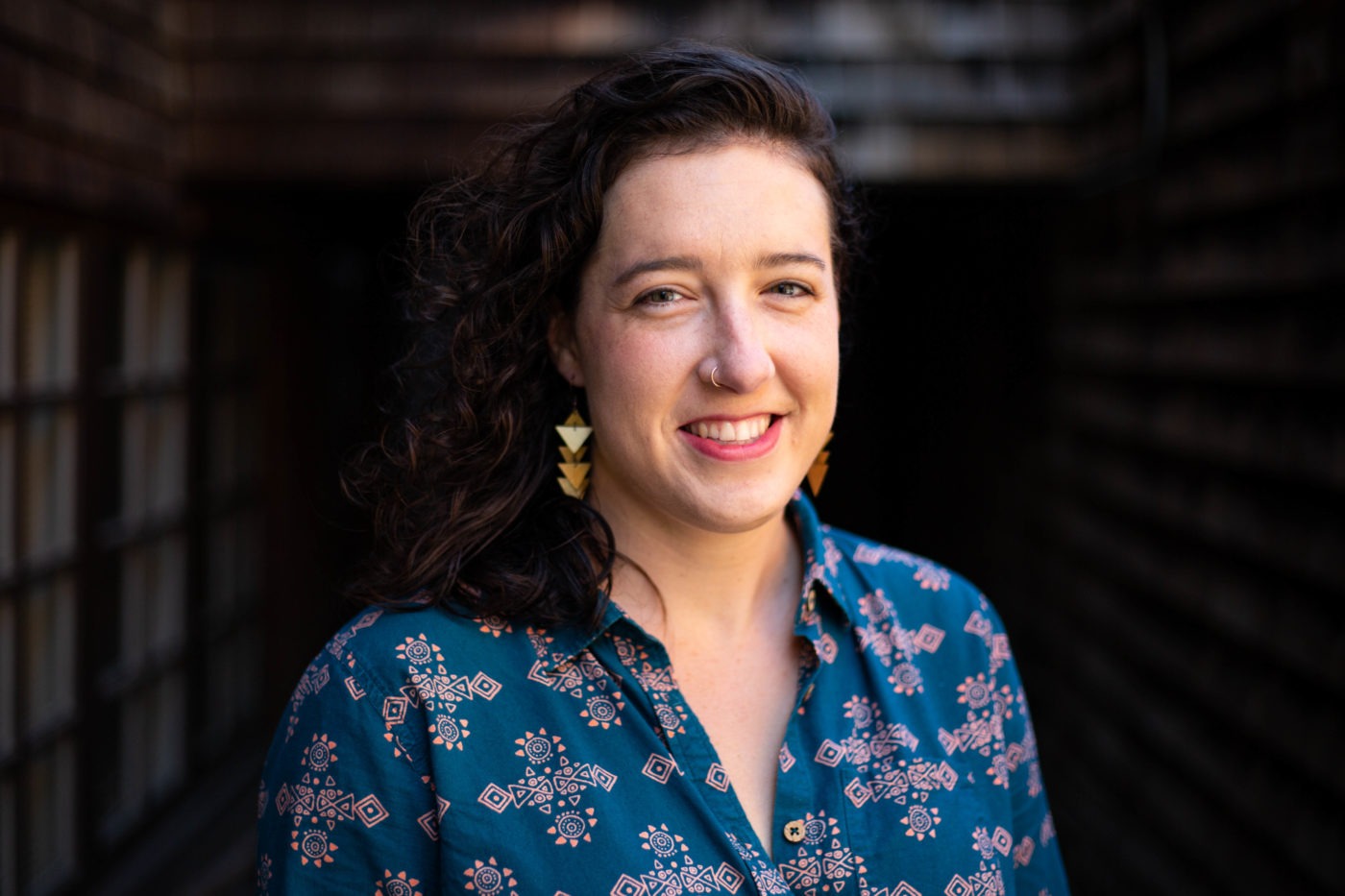 Woman with curly dark hair and geometric earrings smiling at the camera. She is wearing a blue patterned blouse and standing in what appears to be a narrow, outdoor corridor with wooden walls and windows in the background—a perfect setting for her latest Berkeley Journalism assignment.