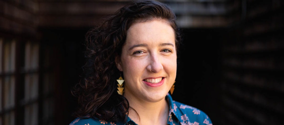 A person with shoulder-length curly hair smiles at the camera. They wear a teal shirt with floral patterns and triangle-shaped earrings, exuding an air of Berkeley Journalism. The background is slightly blurred, indicating a dimly lit corridor or alley.