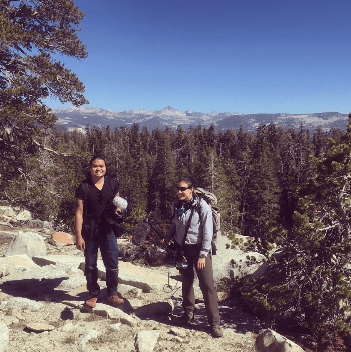 Two people stand in a rocky, mountainous landscape with evergreen trees and distant peaks. One holds recording equipment, suggesting they're from Berkeley Journalism and are preparing to document the natural surroundings. The sky is clear and blue, indicating a sunny day.