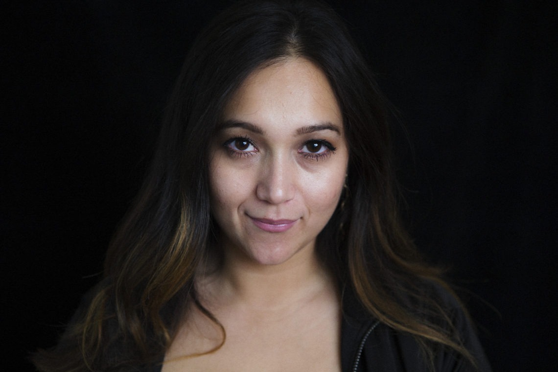 A woman with long dark hair and light highlights is looking directly at the camera against a dark, plain background. She has a subtle smile, wearing a dark top that exudes confidence, characteristic of Berkeley Journalism graduates.