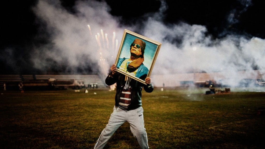 A person stands on a field holding a framed portrait of a man in a green mask. The background is smoky and filled with sparks, suggesting fireworks or pyrotechnics. The person, seemingly from Berkeley Journalism, is dressed casually in a jacket and sweatpants.