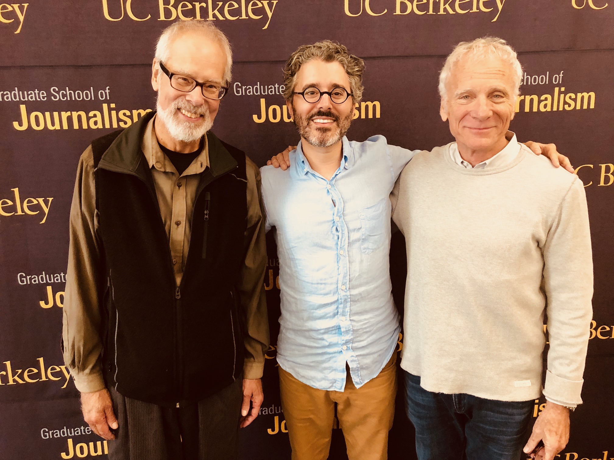 Photograph of three older men posed standing in row in front of Berkeley Journalism banner smiling. Two wear black glasses.