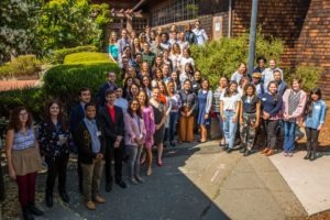 A large group of people pose for a photo outside a building with wood siding at the Berkeley Journalism event. They are smiling, and some are dressed in business-casual attire while others are more casually dressed. The group includes diverse ages and genders, standing in a sunny outdoor setting.