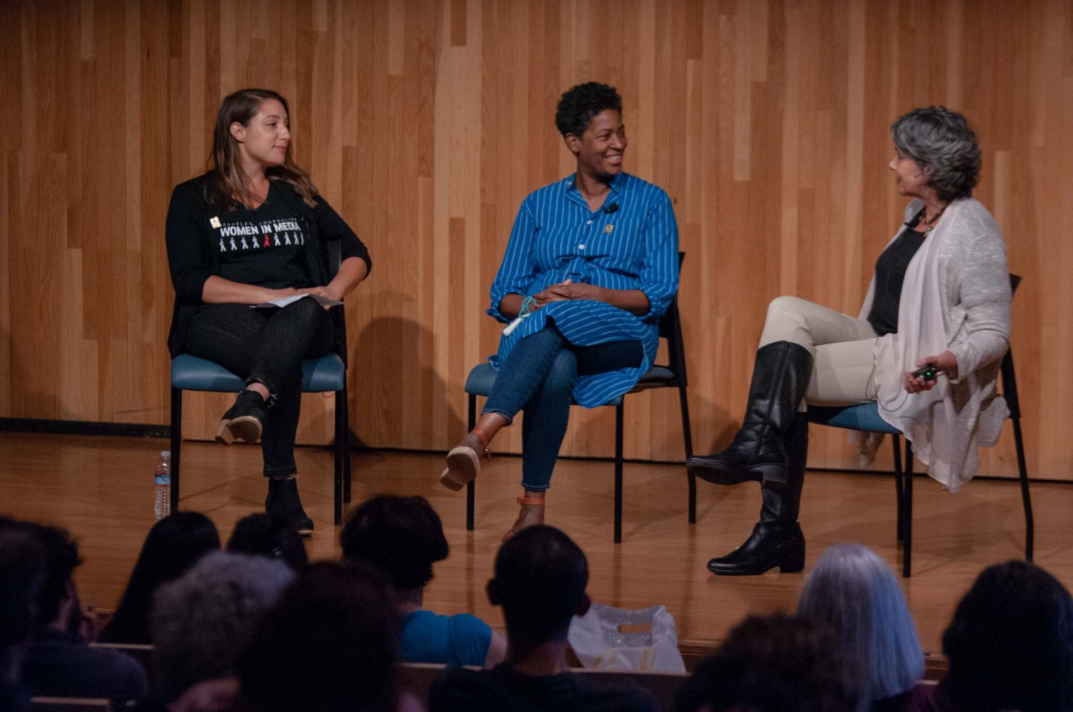 Photo of three women seated on chairs on stage talking and laughing in front of a wood paneled background.