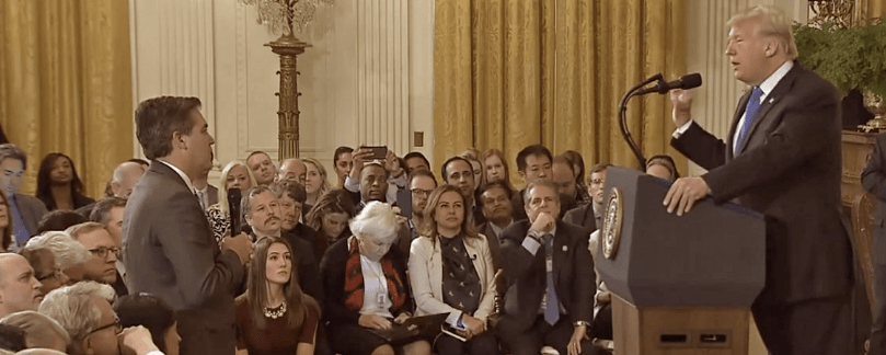 A man stands and speaks into a microphone while another man at a podium responds. The setting appears to be a formal Berkeley Journalism event with numerous people seated and watching attentively. The background features yellow curtains and a chandelier, adding to the distinguished atmosphere.