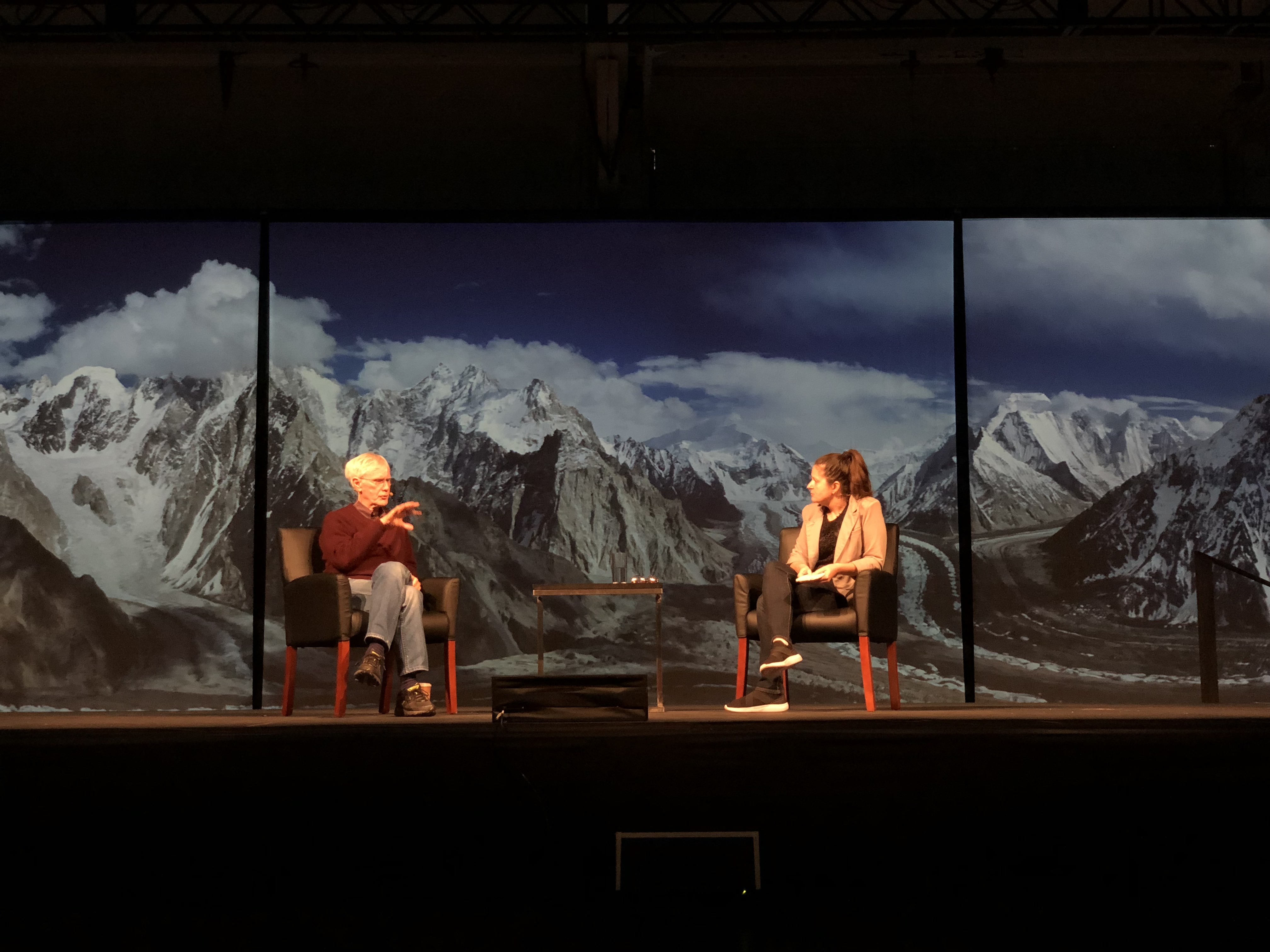 Horizontal photo of an older man with white hair seated on a chair on a stage talking to a woman with brown hair in a ponytail on a stage with large mountain photo in the background.