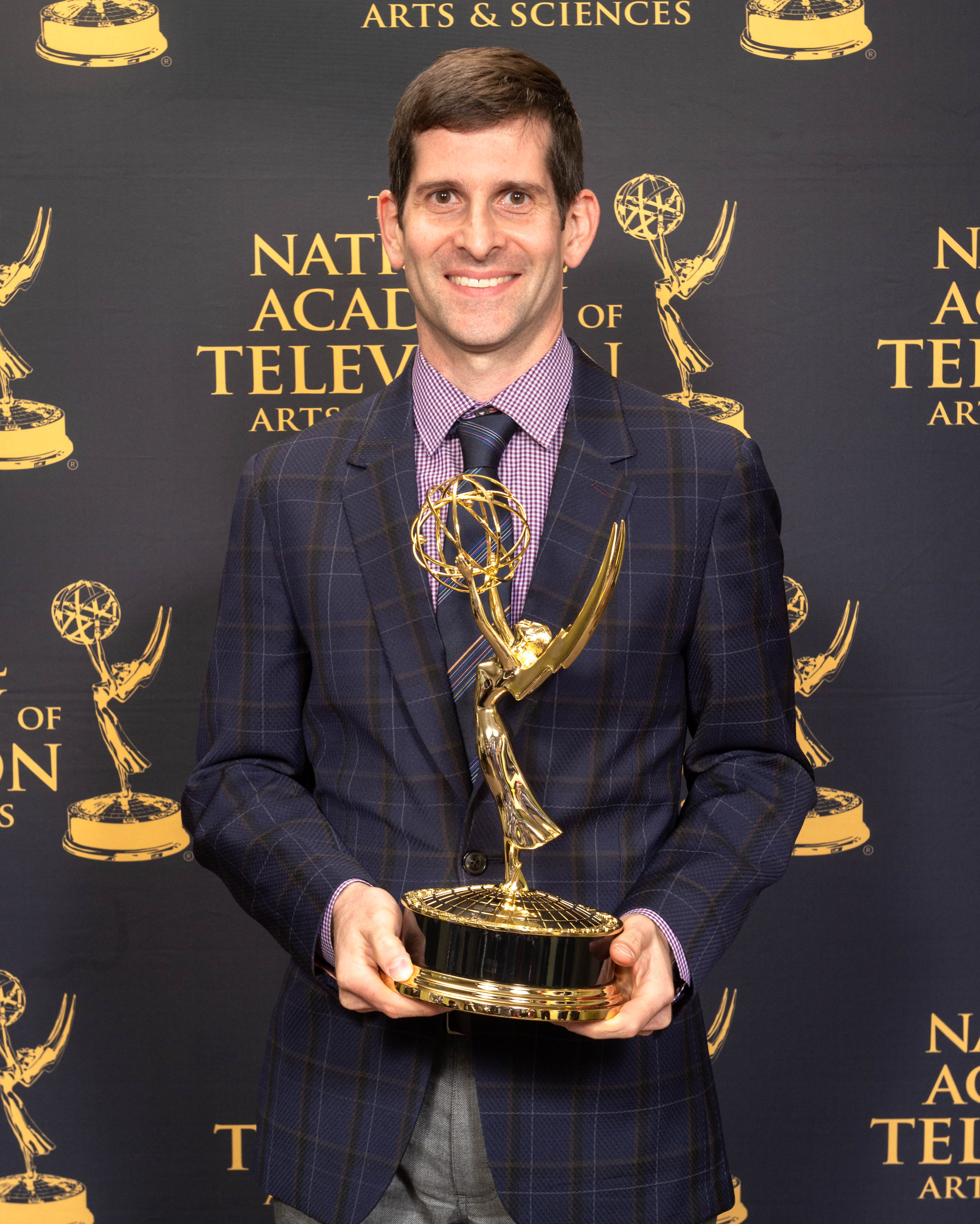 Photo of a man with dark brown closely cropped hair wearing a suit holding an Emmy Award in front of National Academy signage in the background smiling.