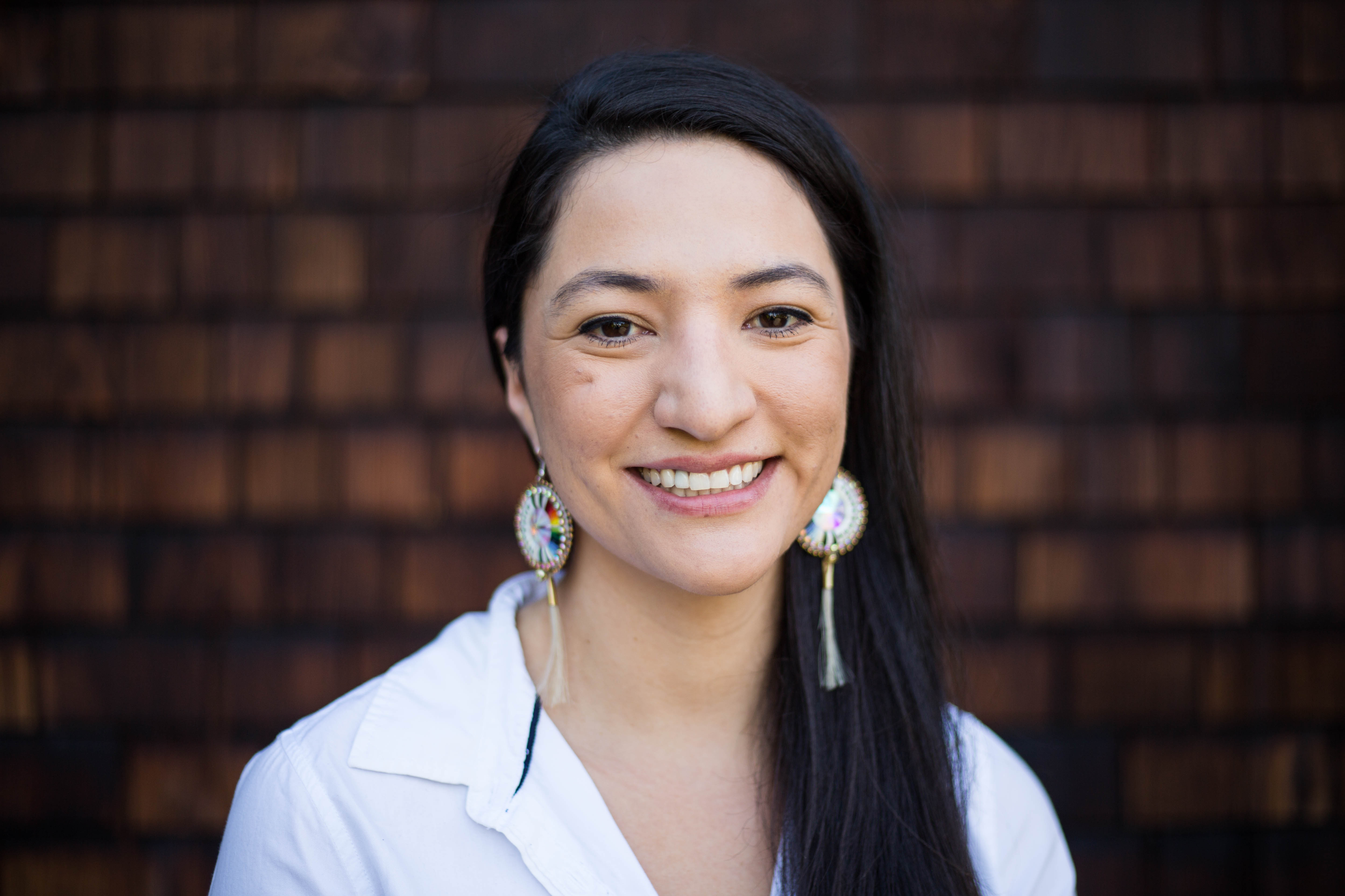 Photograph of a young filmmaker with very long dark straight hair wearing round white tribal earrings and a white blouse in front of a wood shingled background smiling brightly.