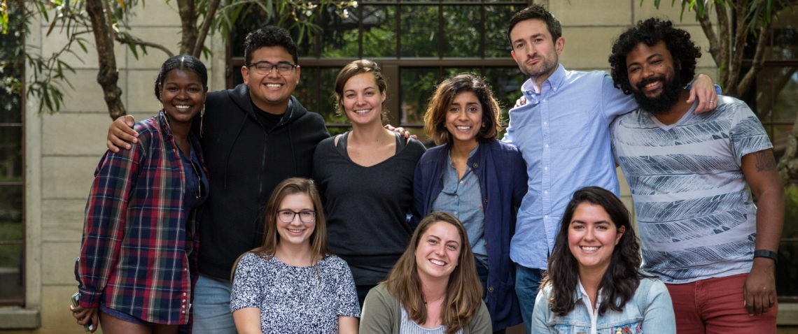 A diverse group of nine people, reflecting a variety of ethnicities, genders, and ages, are standing and sitting together outdoors in front of a building with large windows. They are smiling and appear to be enjoying each other’s company. Trees and greenery are visible in the background. It’s a lively scene reminiscent of the vibrant community at Berkeley Journalism.