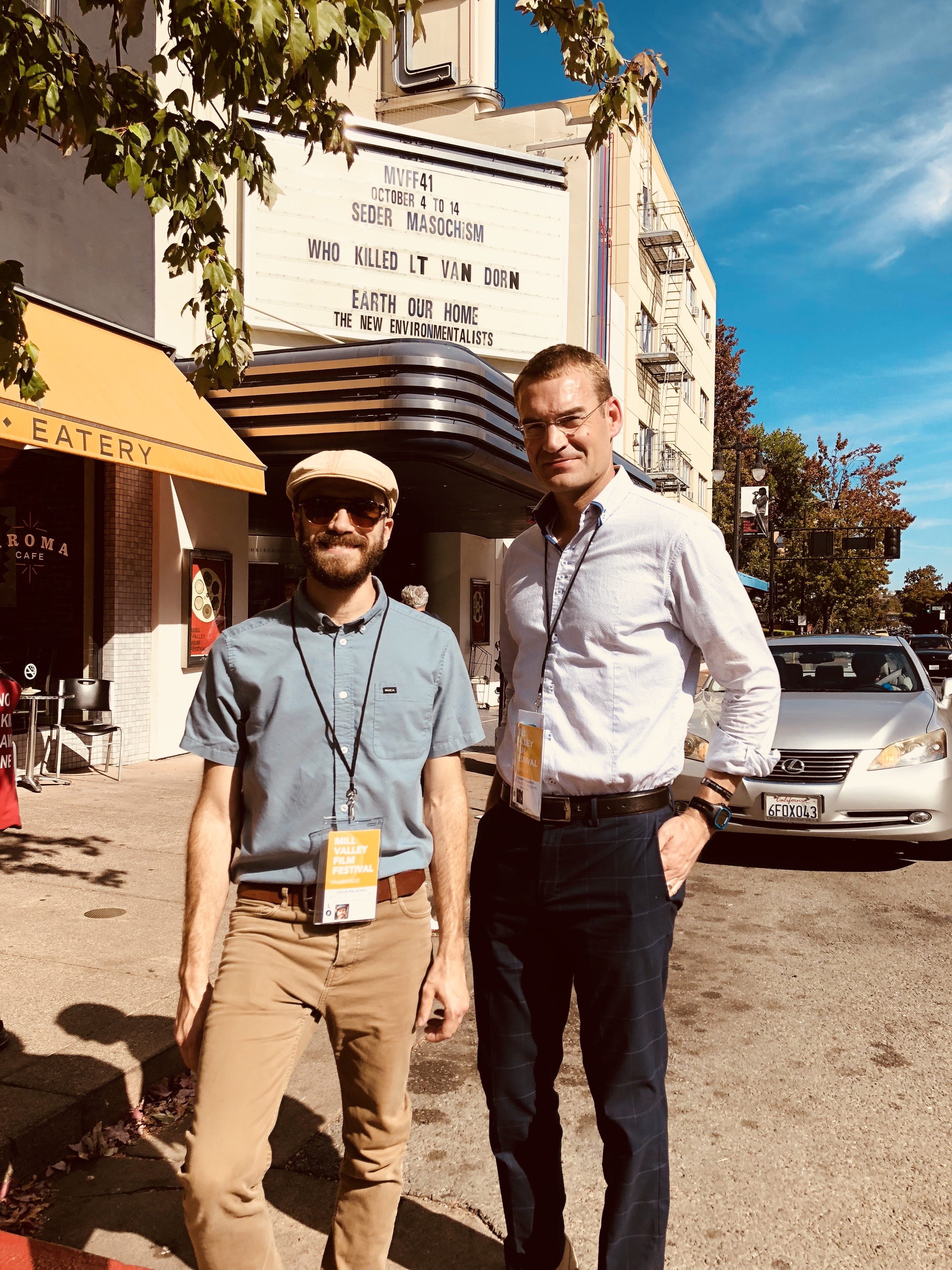 Photograph of two mean standing in front of a theatre outdoors smiling.