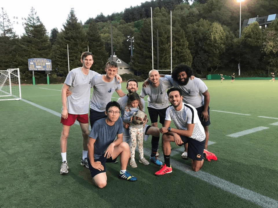 Photo of 5 men and a young girl grouped together on a soccer field wearing athletic gear smiling.