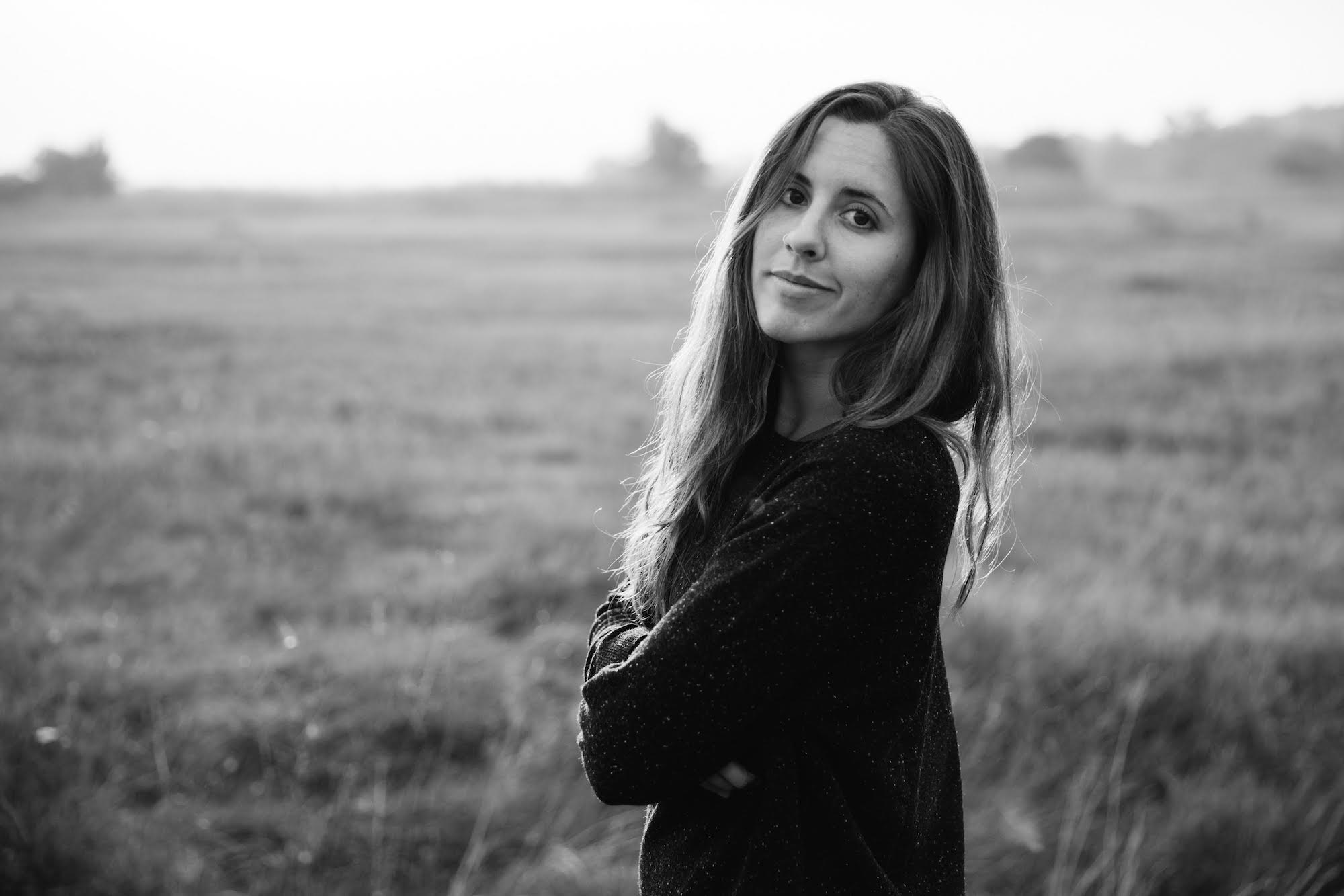 Black-and-white image of a woman with long hair standing in a field. She is wearing a dark sweater, has her arms crossed, and is looking at the camera with a slight smile. The blurred scenic landscape with trees and tall grass evokes the timeless quality often captured by Berkeley Journalism students.