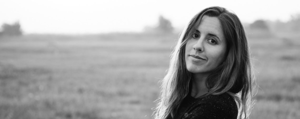 Black and white photo of a woman with long hair standing in a field. She is facing the camera with a slight smile. The background, blurred with grassy terrain and distant trees, evokes the storytelling essence captured at Berkeley Journalism.