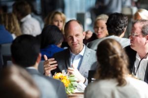 A man in a gray suit is speaking animatedly while seated at a table with several other people in a bright, busy indoor setting. The lively atmosphere feels reminiscent of a Berkeley Journalism workshop. There is a centerpiece with flowers on the table, and the background shows others engaged in conversations.