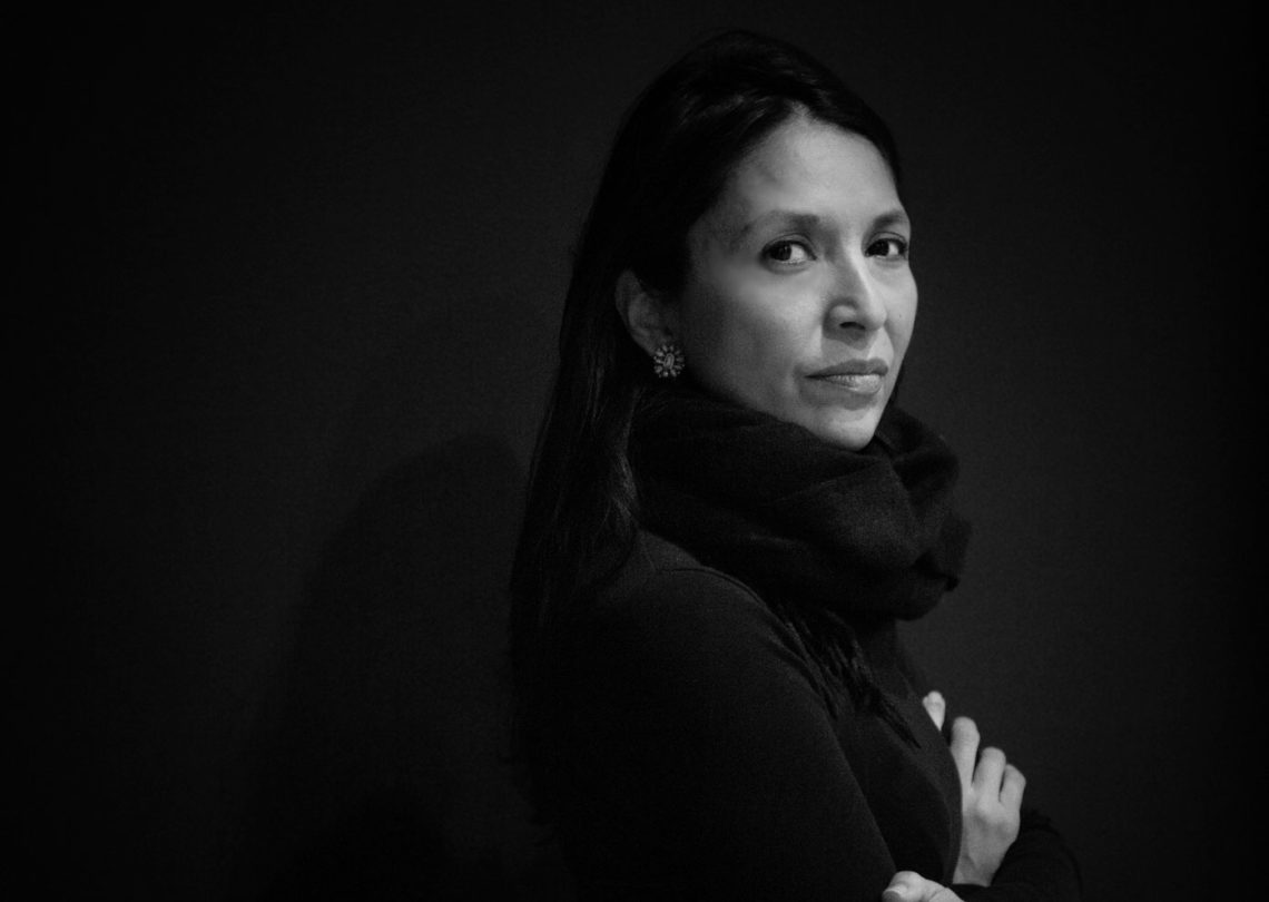 Black and white photo of a woman with long dark hair, wearing a dark outfit and a scarf. She is looking directly at the camera with a serious expression, her arms folded. The plain, dark background enhances the focus on her face, capturing the contemplative mood of Berkeley Journalism.