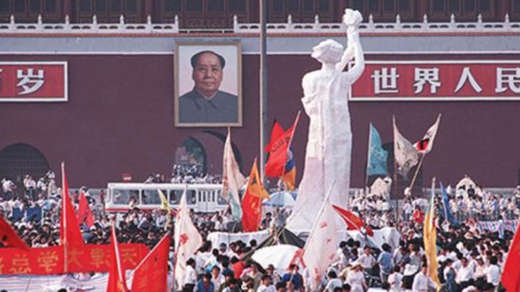 A large gathering of people with various colorful flags converges around a tall, white statue in a public square. A significant portrait of a man hangs on the red wall of a building in the background, reminiscent of scenes often captured by Berkeley Journalism students.