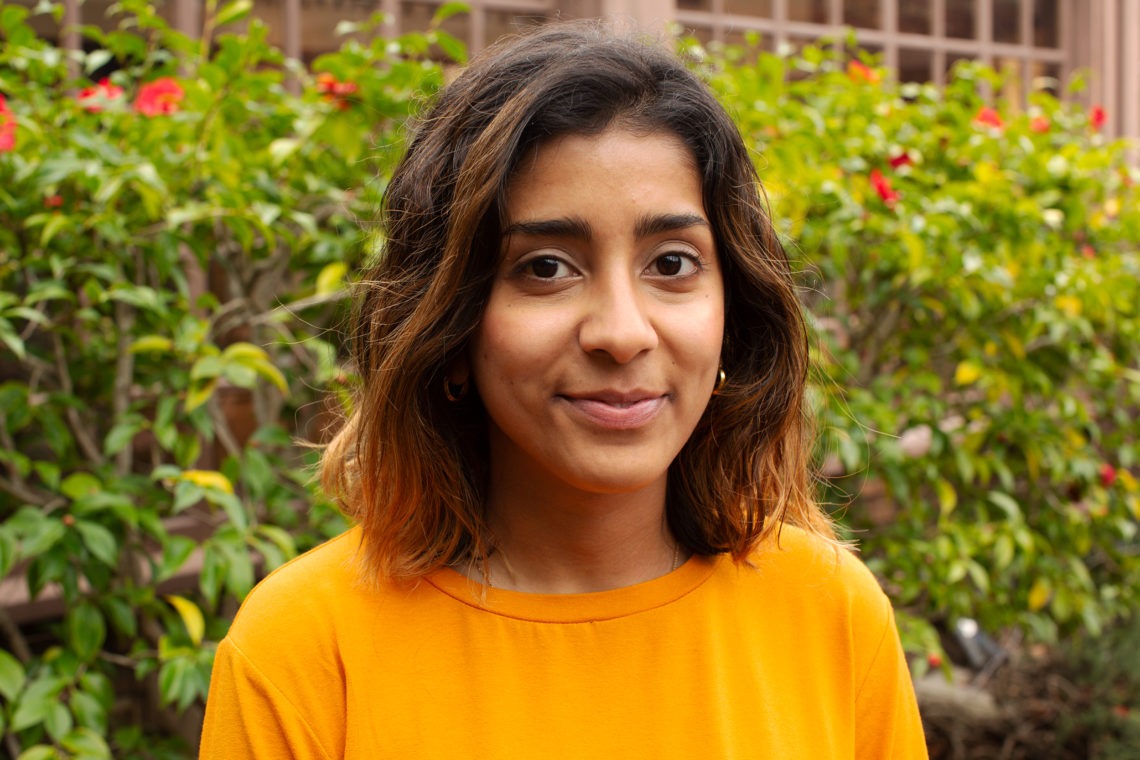 A woman with shoulder-length brown hair is wearing an orange shirt and standing in front of lush green bushes with red flowers. She is looking into the camera, smiling gently, exuding a warmth that reflects her dedication to stories from Berkeley Journalism.