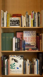 A wooden bookshelf filled with various books of different sizes and colors evokes the spirit of Berkeley Journalism. Notable titles include "The Americans" by Robert Frank, "Observations" by Richard Avedon, and "One decisive moment." The books are neatly arranged in multiple rows.