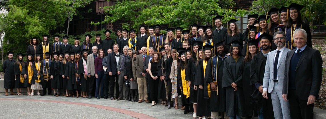 Class of 2019 Berkeley Journalism graduates in black caps and gowns pose outdoors with faculty members and other participants. Some graduates wear yellow sashes. Trees and a building can be seen in the background. Everyone appears happy and celebratory.