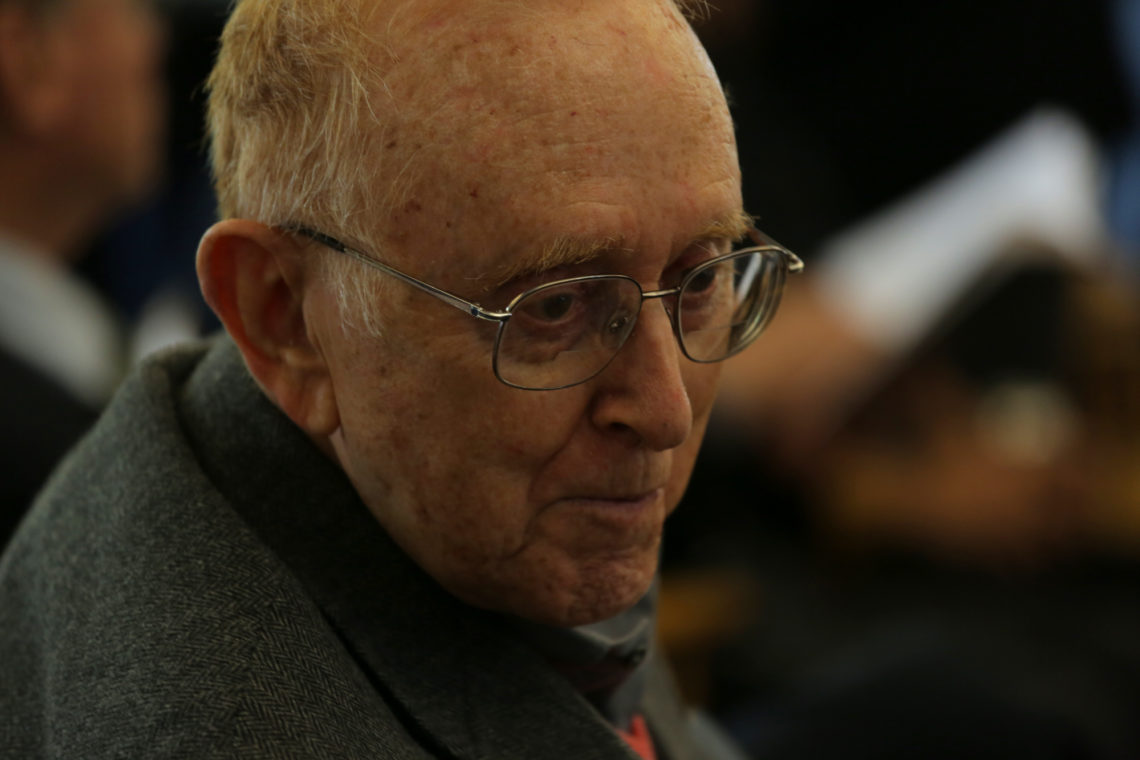 A close up photo of an elderly man looking into the distance. He is smiling, wearing a suit, and glasses. 