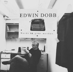 A black and white photograph shows a person sitting in an office, reading a paper. On the wall behind them is a large sign featuring an old-fashioned typewriter with text that reads, 