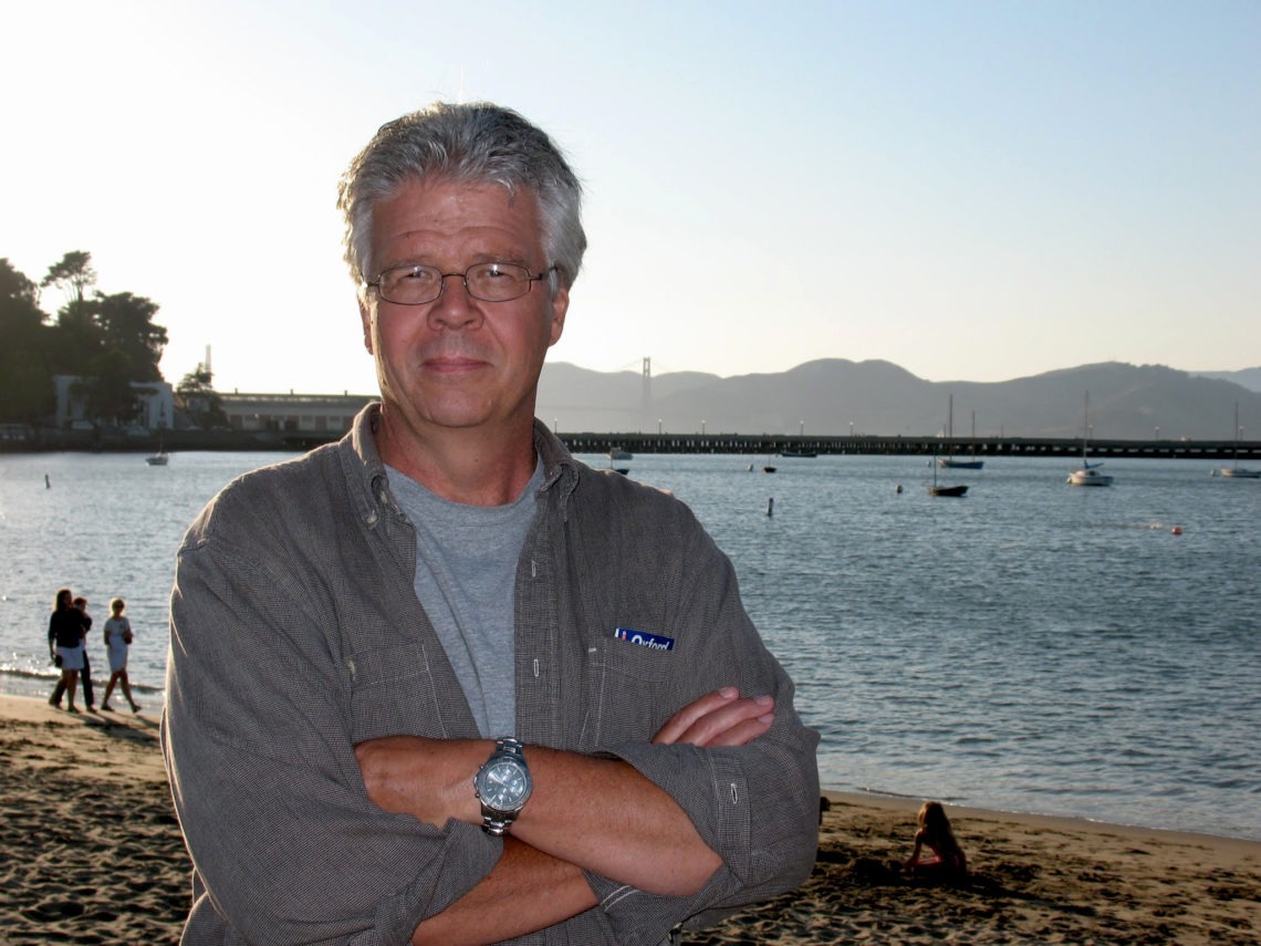 A man with gray hair and glasses stands on a beach with his arms crossed, wearing a gray button-up shirt over a T-shirt. The background features calm water, several boats, a bridge, distant mountains, and a few people walking on the shore—an ideal spot for inspiration in Berkeley Journalism.