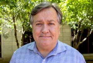 Portrait of a middle-aged person with graying hair, wearing a blue plaid shirt, standing outdoors in front of a background of green trees and a beige-colored building. The person, embodying the spirit of Berkeley Journalism, is looking directly at the camera with a neutral expression.