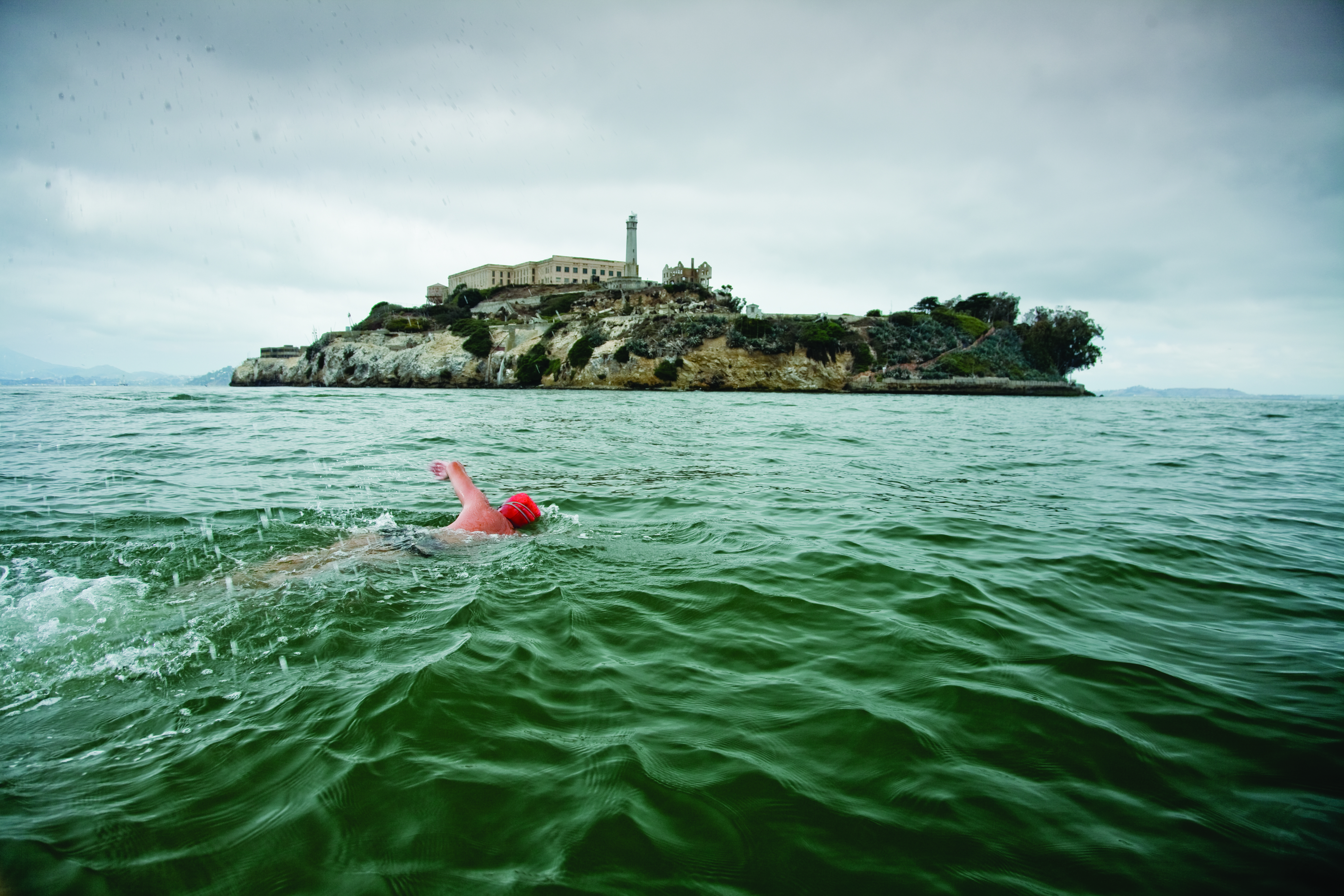 A man in a red swim cap is swimming to Alcatraz island. It is cloudy outside and the water has a strong current. 