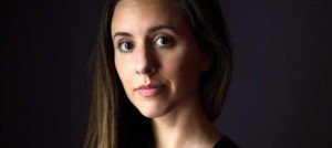 A close-up photograph of a person with long brown hair and brown eyes, looking directly at the camera with a neutral expression. The dark background softly contrasts with their face, capturing a moment that echoes the storytelling essence found in Berkeley Journalism.