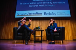 Two people are seated on a stage in armchairs, engaged in an animated discussion. Behind them is a screen with a Berkeley Journalism banner and details about the conversation topic, which includes the "Green New Deal." Both individuals from Berkeley Journalism are gesturing expressively.