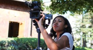 A woman stands outdoors operating a video camera mounted on a tripod. She looks intently at the camera's viewfinder, with trees and a building in the background. As a Berkeley Journalism student, she appears focused on capturing the footage accurately.