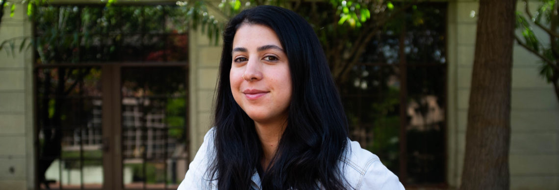 A person with long dark hair and a slight smile stands outdoors in front of a building with large windows and greenery at Berkeley Journalism. The person is wearing a light-colored shirt and looking directly at the camera.