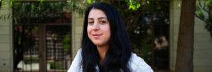 A person with long dark hair and a slight smile stands outdoors in front of a building with large windows and greenery at Berkeley Journalism. The person is wearing a light-colored shirt and looking directly at the camera.