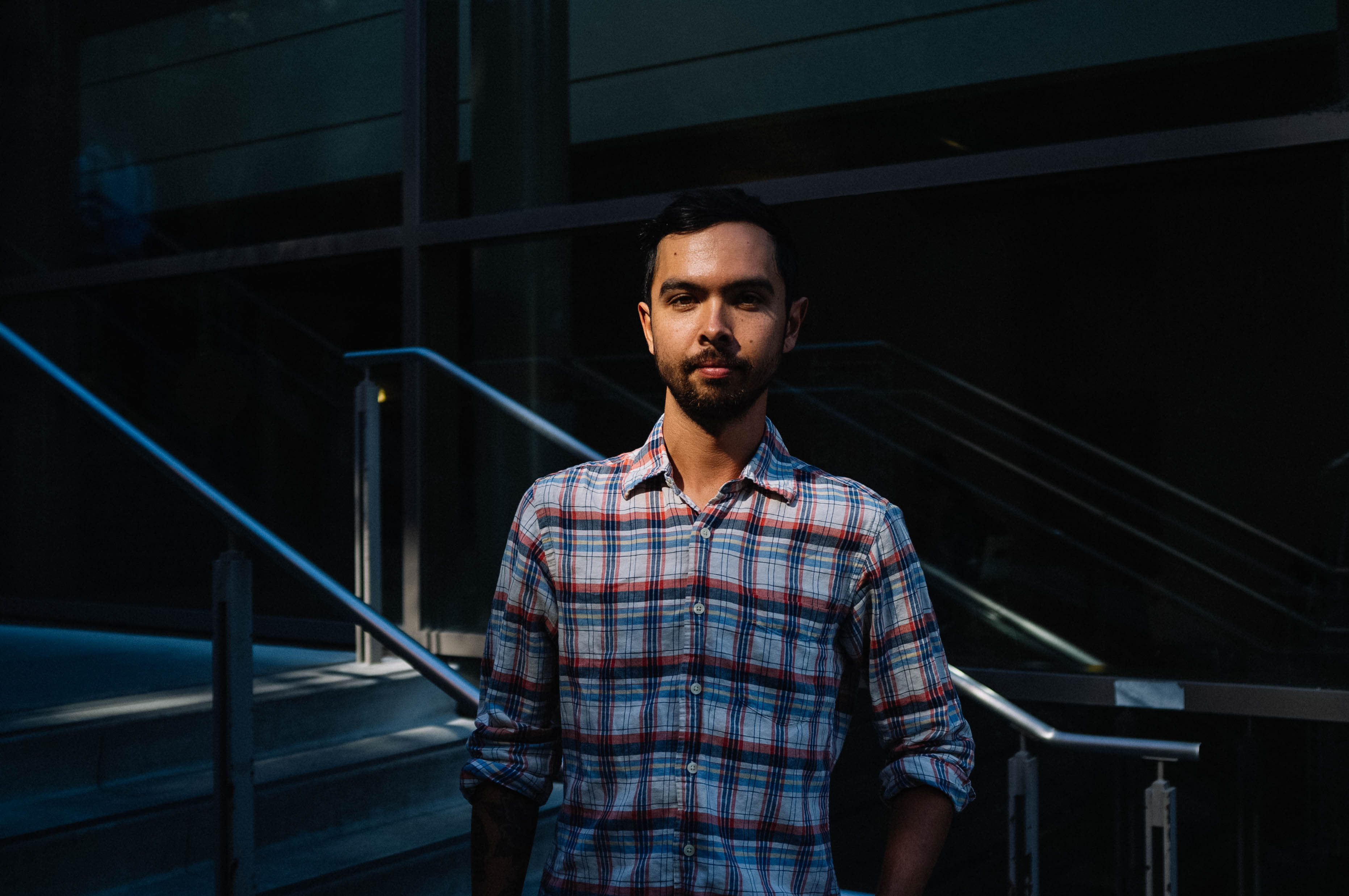 A dark filtered photo of a man standing in front of stairs and guard rails. he has a slight smile and is wearing a plaid shirt.