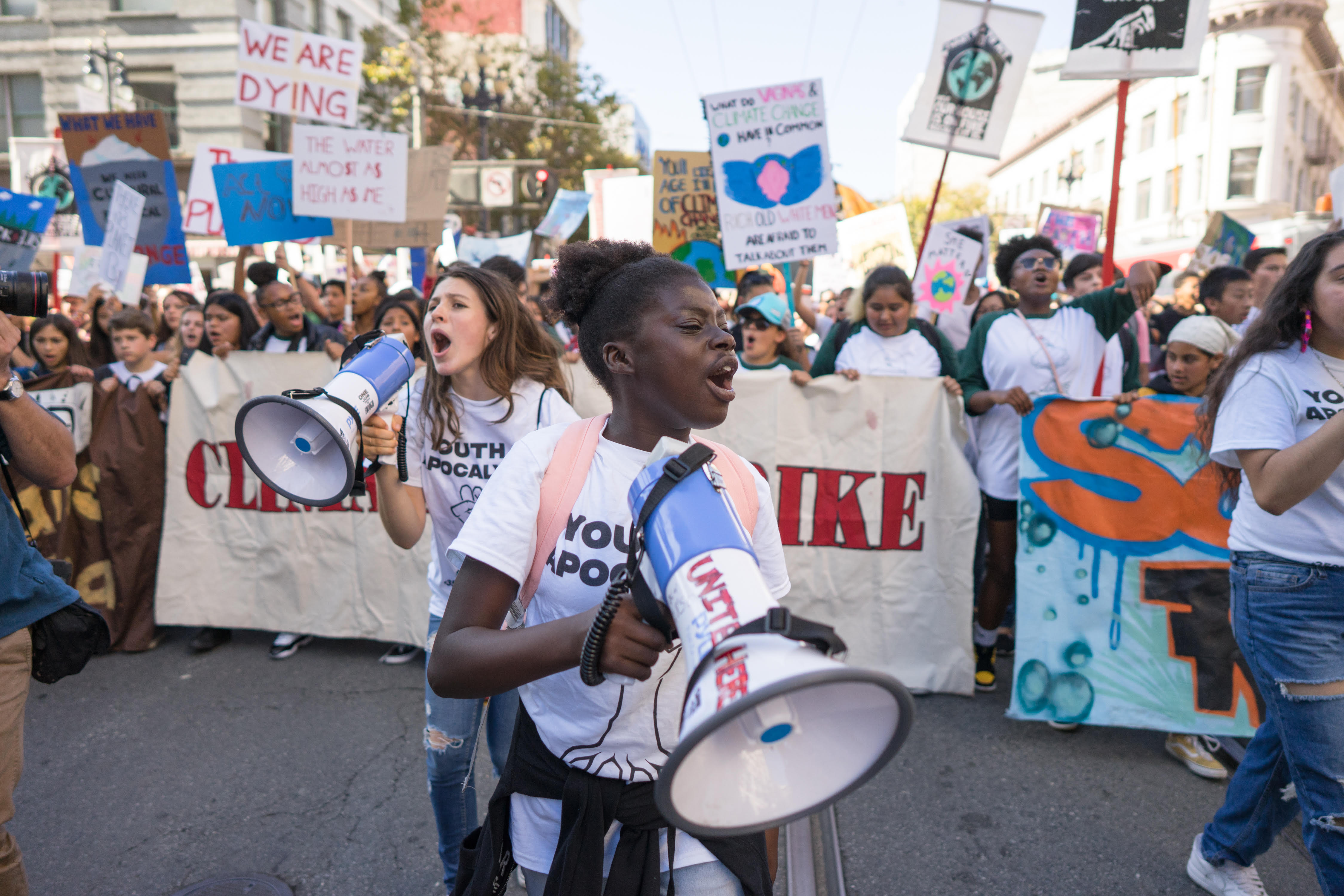 Two kids holding megaphones in the street protesting with a massive group following behind. They are holding large banners and picket signs. 