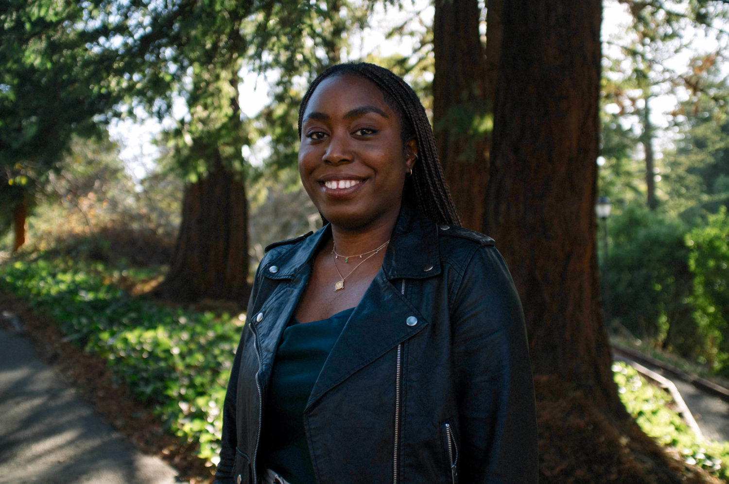 A woman with long braids wearing a leather jackets stands outside for a portrait. She is surrounded by trees and the sun is peering through.