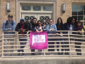 A group of people standing on a balcony hold a pink banner. The banner says "Full Frame".