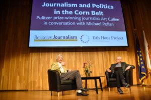 Two men sit in chairs on a stage engaged in conversation. Behind them, a screen displays the title "Journalism and Politics in the Corn Belt," featuring Pulitzer-winning journalist Art Cullen interviewing Michael Pollan. The logos for Berkeley Journalism and 11th Hour Project are also visible.