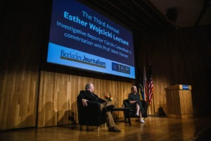 A man and a woman are seated on a stage in front of a large screen displaying "The Third Annual Esther Wojcicki Lecture," hosted by Berkeley Journalism and the Taube Family Foundation. The woman holds a microphone, and the podium proudly displays the same text.