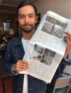 A man with a beard and mustache, dressed in a denim jacket and white t-shirt, stands indoors holding up a newspaper. The background, reminiscent of a Berkeley Journalism setting, shows people seated at tables, a framed picture on the wall, and a guitar hanging up.