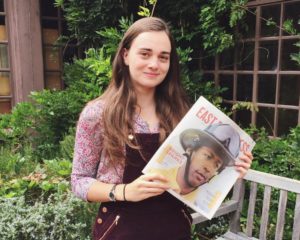 A person with long hair, wearing a patterned shirt and maroon overalls, stands outside holding up a Berkeley Journalism magazine titled "EAST" with a cover featuring a person in a helmet. They are in a garden with lush green plants and a wooden bench in the background.