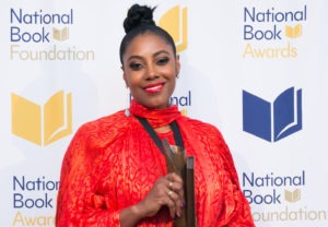 A woman in a vibrant red-orange outfit, a graduate of Berkeley Journalism, holds a National Book Award trophy while smiling at the camera. The backdrop features the logos of the "National Book Foundation" and "National Book Awards".