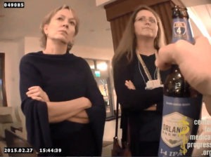 Two women stand indoors with their arms crossed, facing a person holding a bottle of Orlando brew. The timestamp on the image reads "2015.02.27 15:44:39" and the ID number is "049085." Notably, a logo at the bottom right corner reads "Center for Medical Progress," hinting at possible Berkeley Journalism coverage.