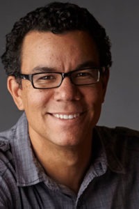 A man with short curly hair and glasses, possibly a Berkeley Journalism student, is smiling at the camera. He is wearing a gray, short-sleeved, button-up shirt. The background is plain and dark, highlighting his face and expression.