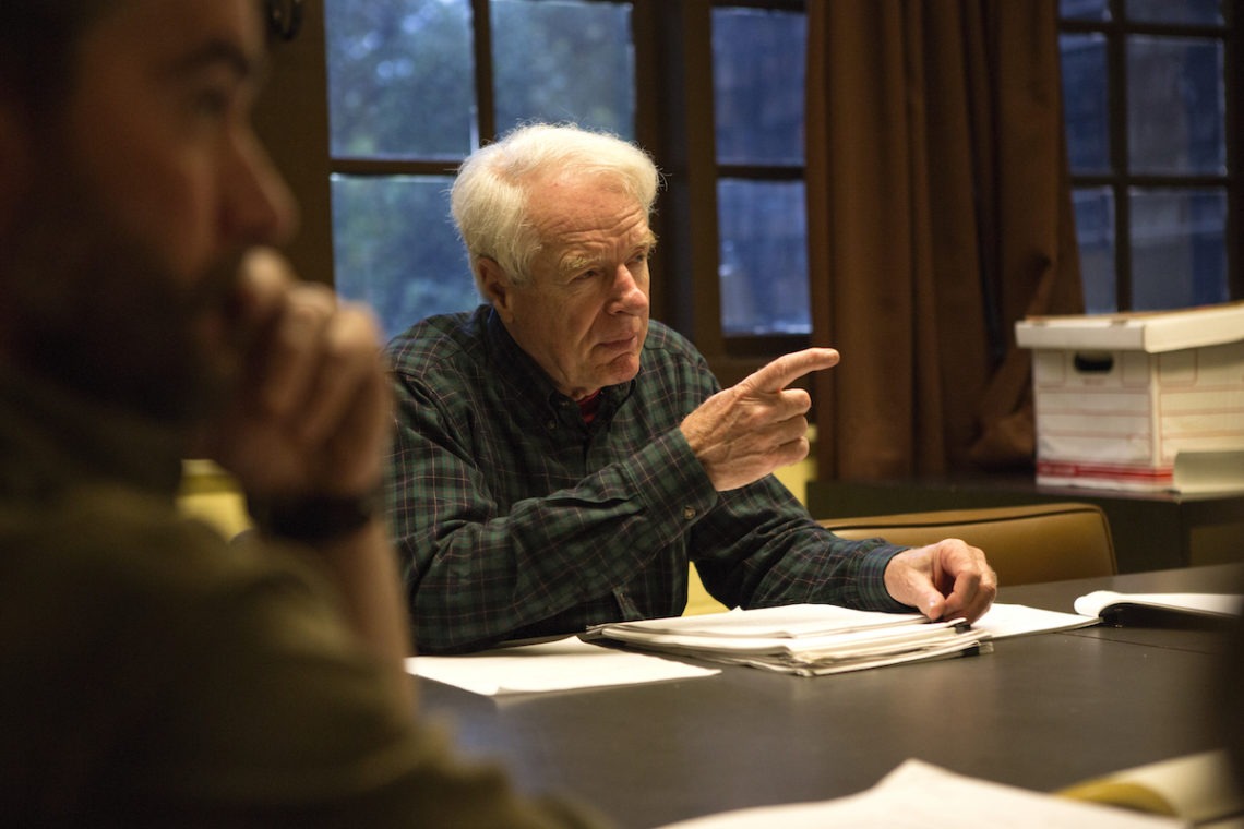 An elderly man with white hair and wearing a green plaid shirt gestures with his hand while speaking at a table, surrounded by papers. He sits near a window, deep in discussion with another person in the foreground, partially out of focus. The setting has the thoughtful ambiance of a Berkeley Journalism seminar.